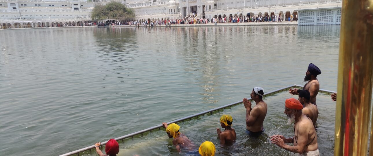 Devotees bathing in the sacred Sarovar at the Golden Temple in Amritsar, with the temple reflected in the calm water during the daytime.